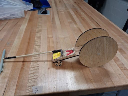 Mousetrap car with a pair of large plywood rear wheels on a butcher-block bench