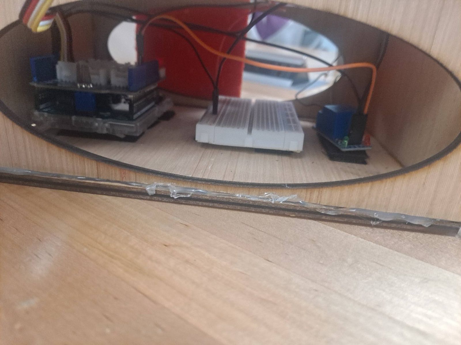 Close-up through an oval cutout of the breadboard and relay wiring inside the control box
