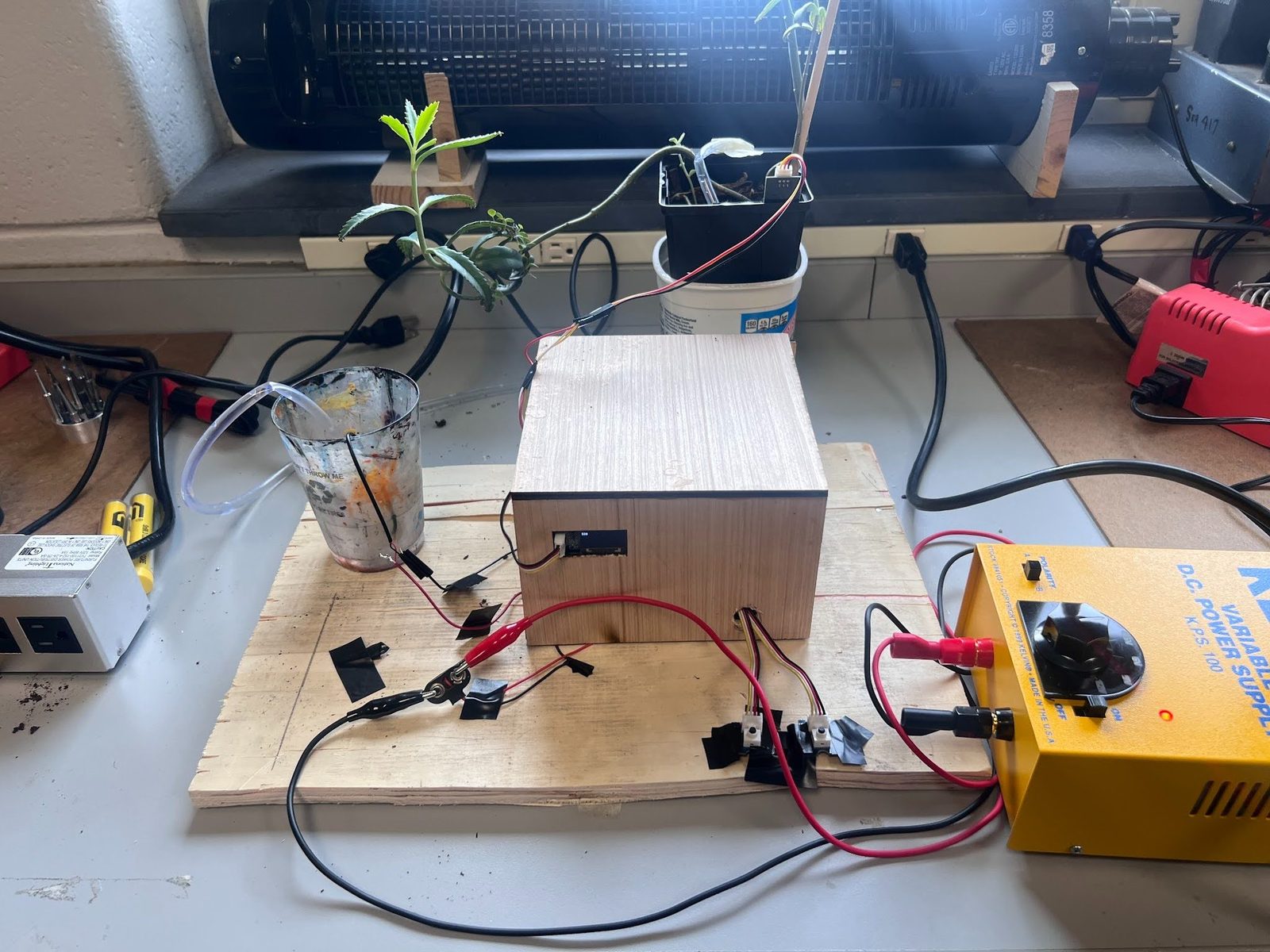 Full plant watering setup on a workbench with potted plants, tubing, and a variable DC power supply
