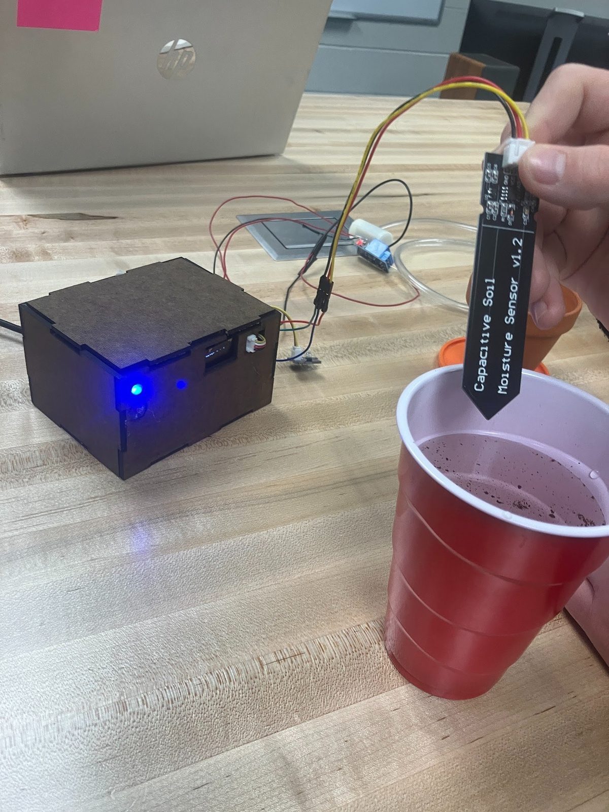 Hand holding a capacitive soil moisture sensor over a cup of soil, with the laser-cut control box lit up in the background