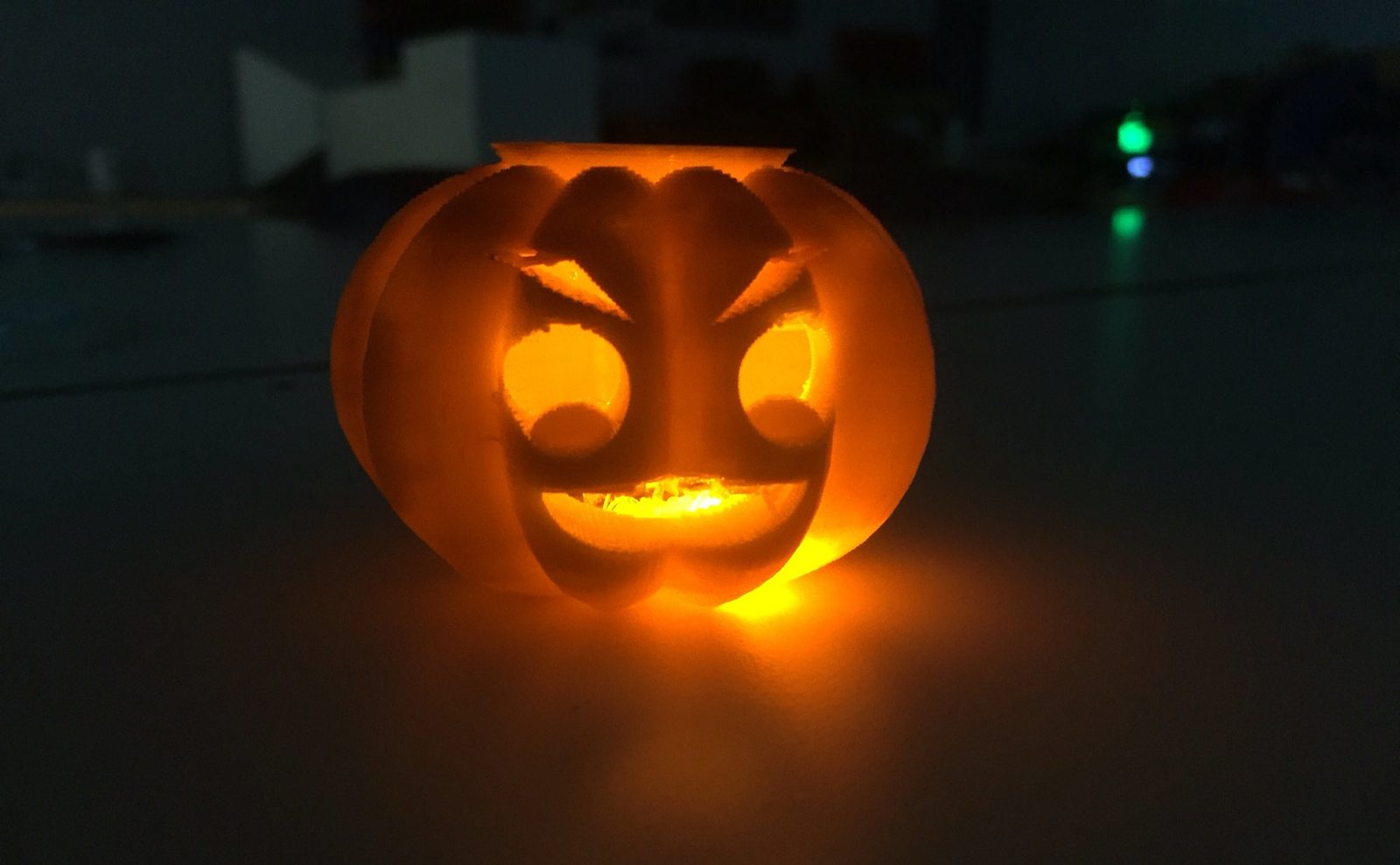 Close-up of a single glowing 3D printed jack-o-lantern with a grinning face, lit from within in a dark room