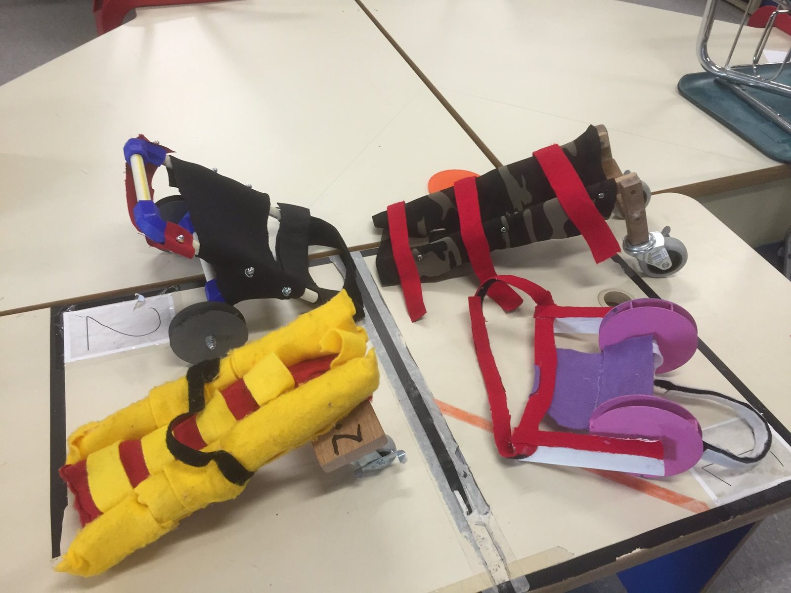 Several fabric-wrapped prototype wheelchairs for injured dogs, built on wheeled wooden frames, numbered as student design entries
