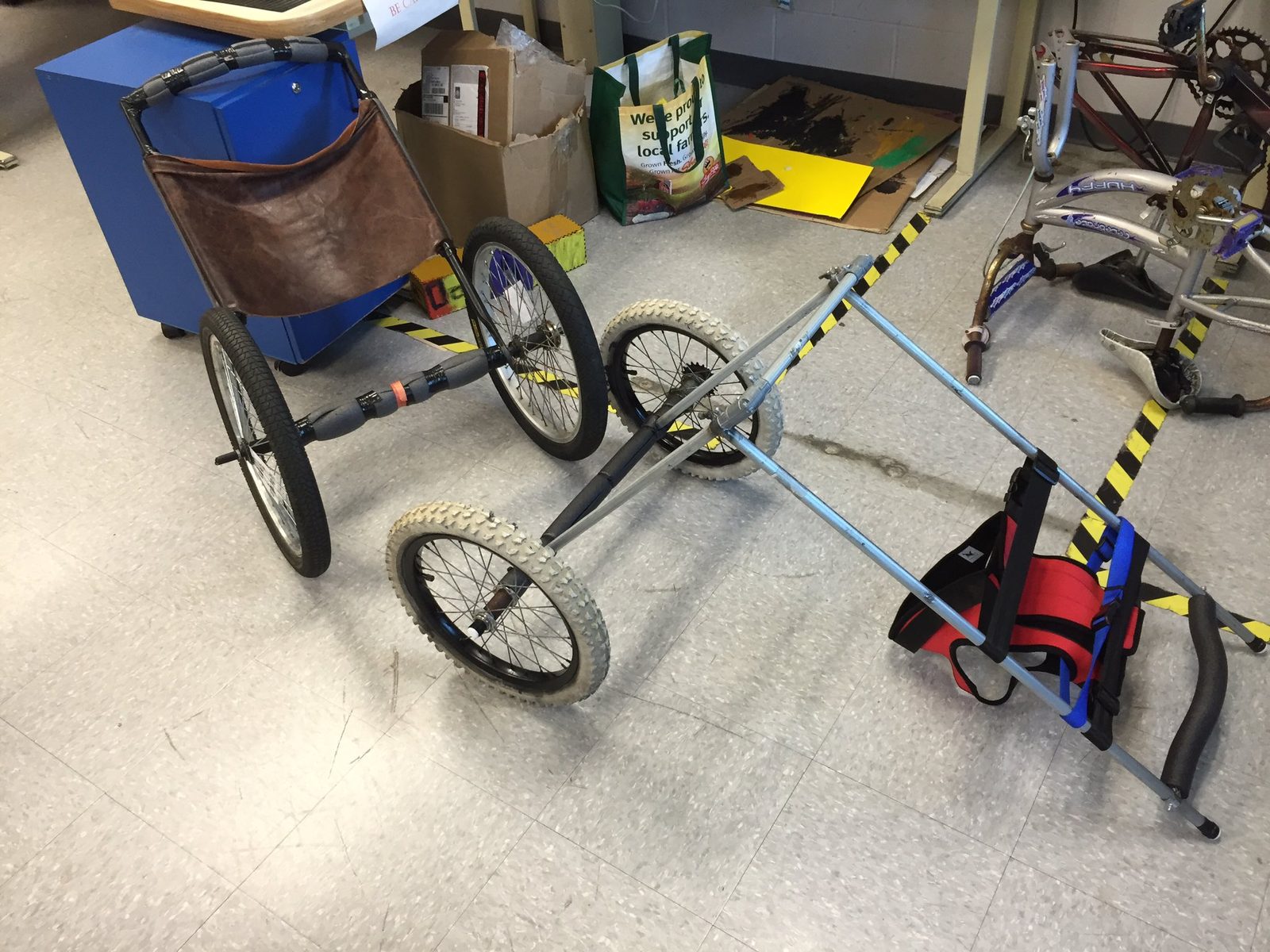 A larger prototype dog wheelchair welded from bike wheels and tubing, with a padded harness, in the classroom
