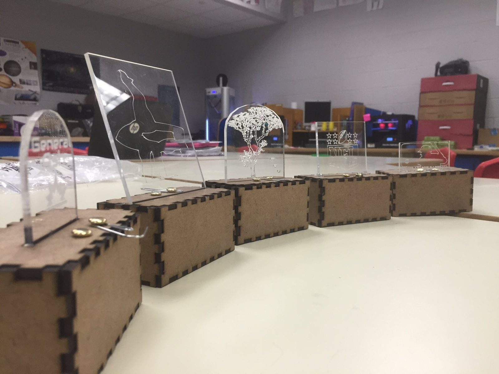 Row of finished light boxes with laser-cut wooden bases and interchangeable acrylic panels engraved with different designs, including an HP logo and an NFL logo