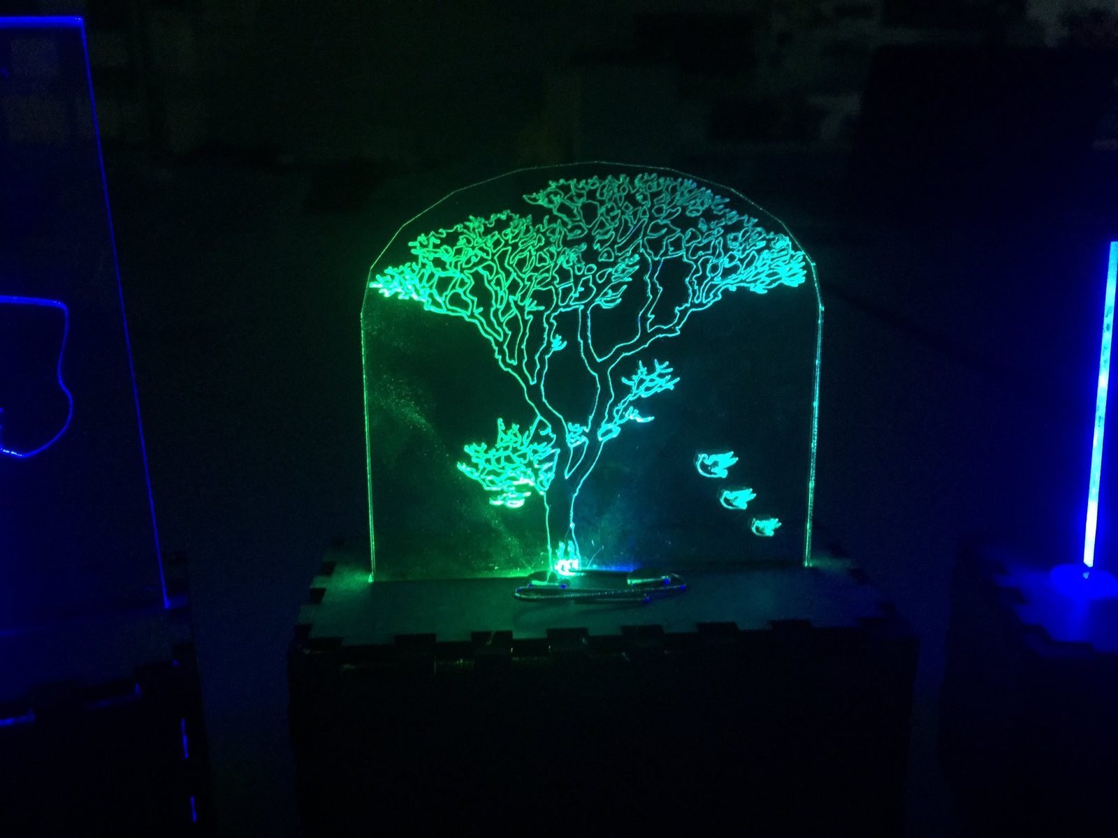 Edge-lit acrylic panel engraved with a tree and birds, glowing green in a dark room
