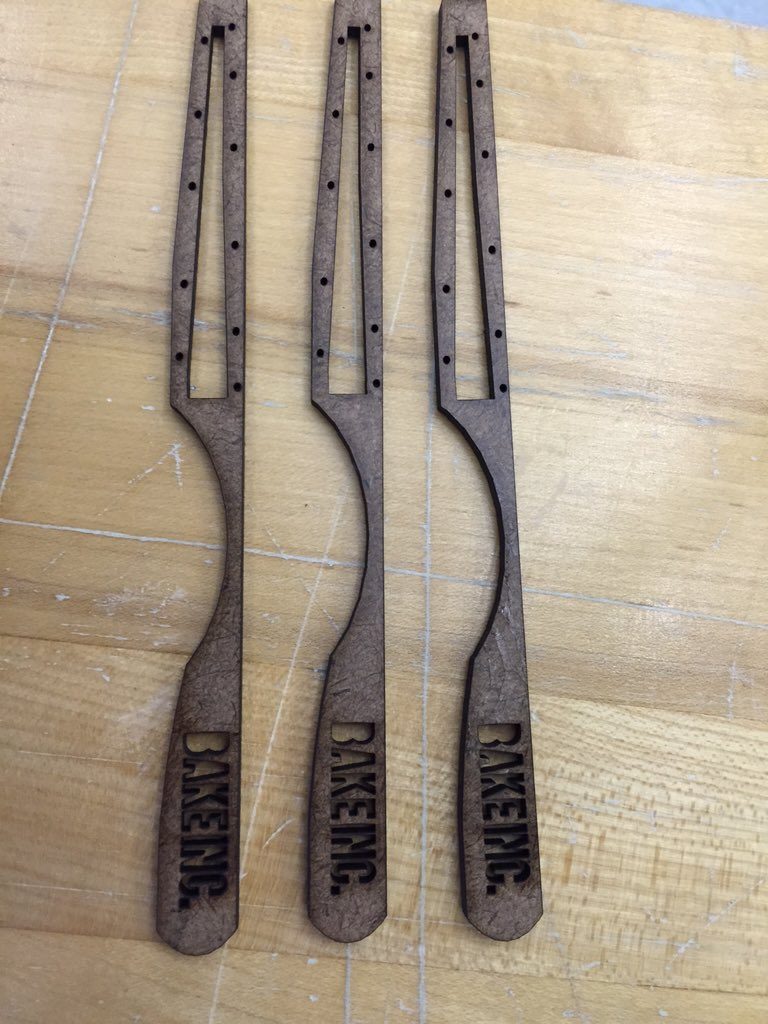 Three laser-cut wooden sunglasses arms engraved with the BAKE INC. logo, laid out on a workbench