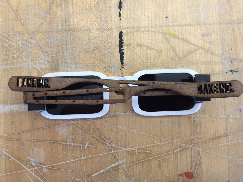 Top-down view of the finished sunglasses showing both engraved BAKE INC. arms folded open