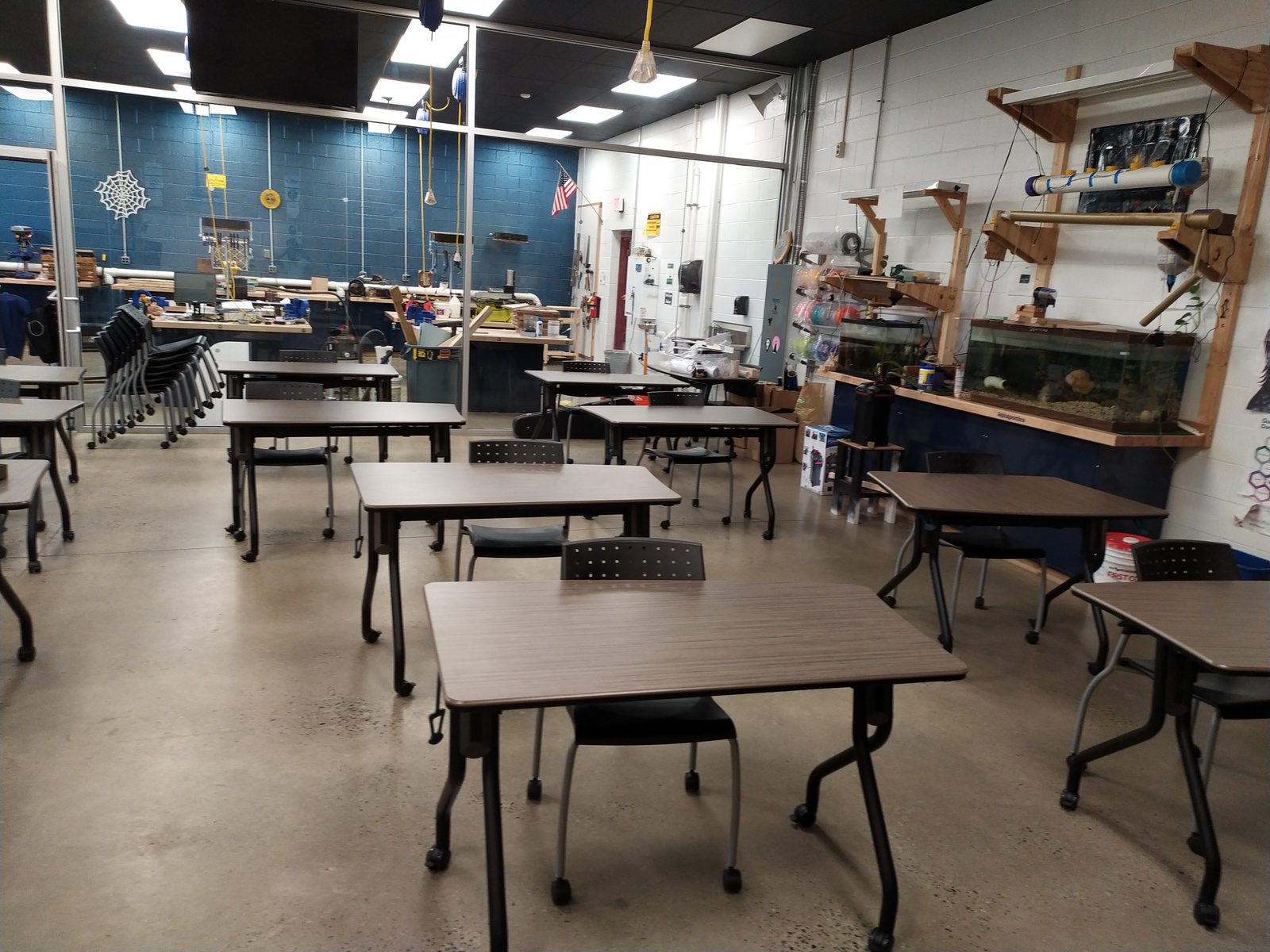 Rows of classroom tables and chairs with the aquaponics tank and fabrication benches visible along the back wall