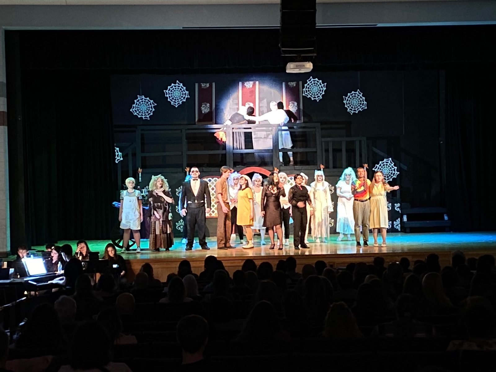 Full cast on stage in costume and makeup for The Addams Family, performing in front of the built set