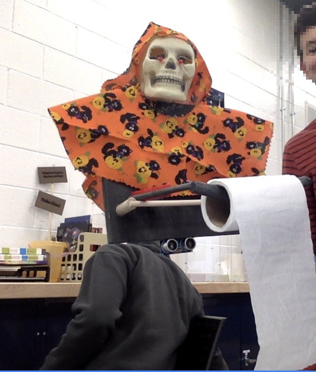 Skeleton animatronic wrapped in orange pumpkin-print fabric, rigged as a toilet paper roll dispenser gag