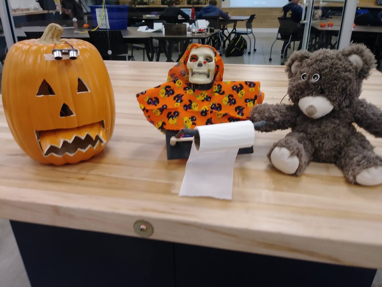 Three Halloween animatronics together on a table: a sensor-eyed jack-o'-lantern, a robed skeleton, and a teddy bear with sensor eyes