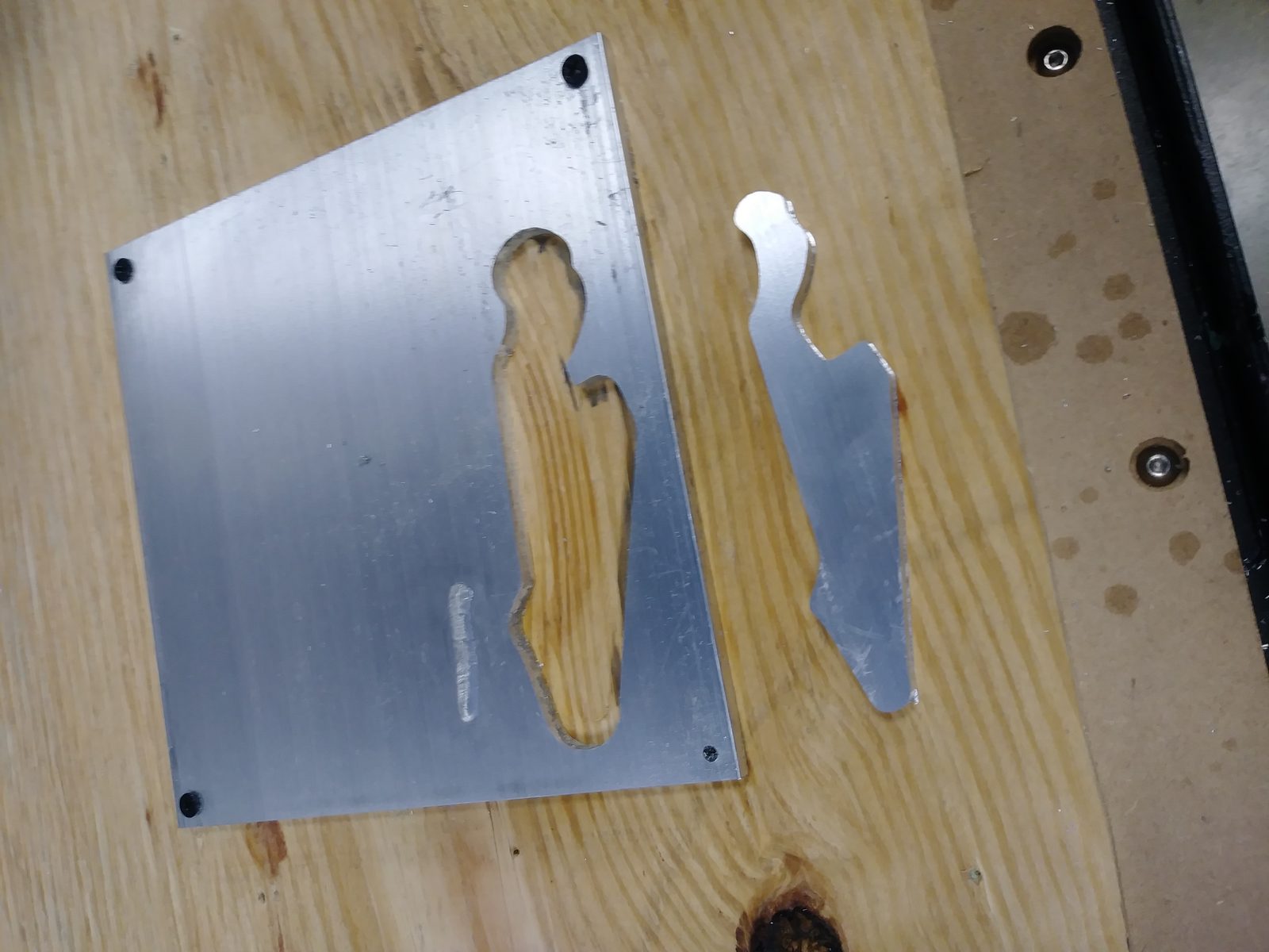 CNC-milled aluminum sheet with a shape cut out, next to the cut piece on a wooden spoil board