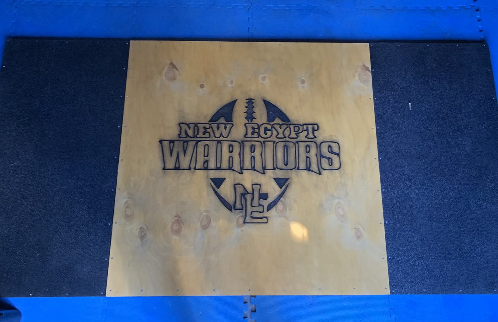 Larger laser-etched wooden panel of the New Egypt Warriors mascot logo mounted on the floor