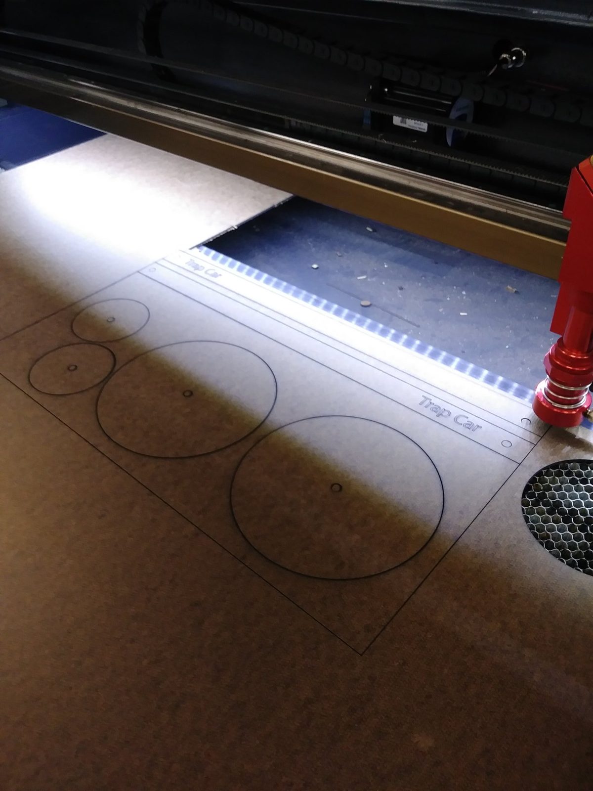 Laser cutter mid-cut on a wooden mousetrap car wheel piece labeled Trap Car