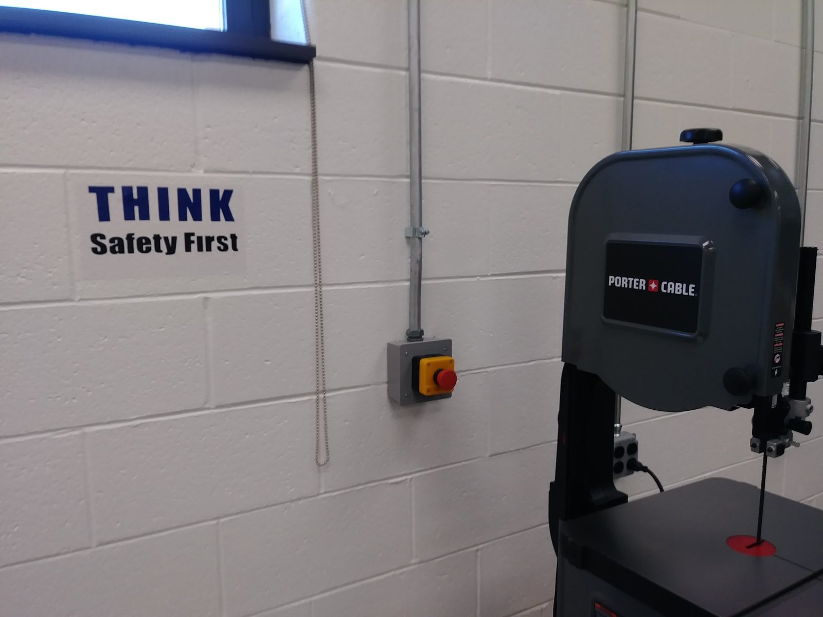 A Think Safety First sign and emergency stop button next to a Porter-Cable band saw in the finished lab
