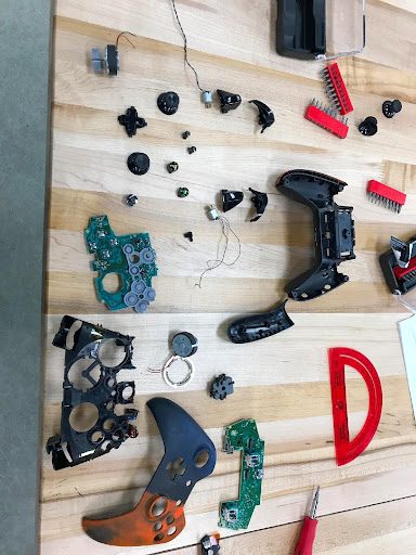 Fully disassembled game controllers laid out on a workbench, with circuit boards, buttons, and triggers separated