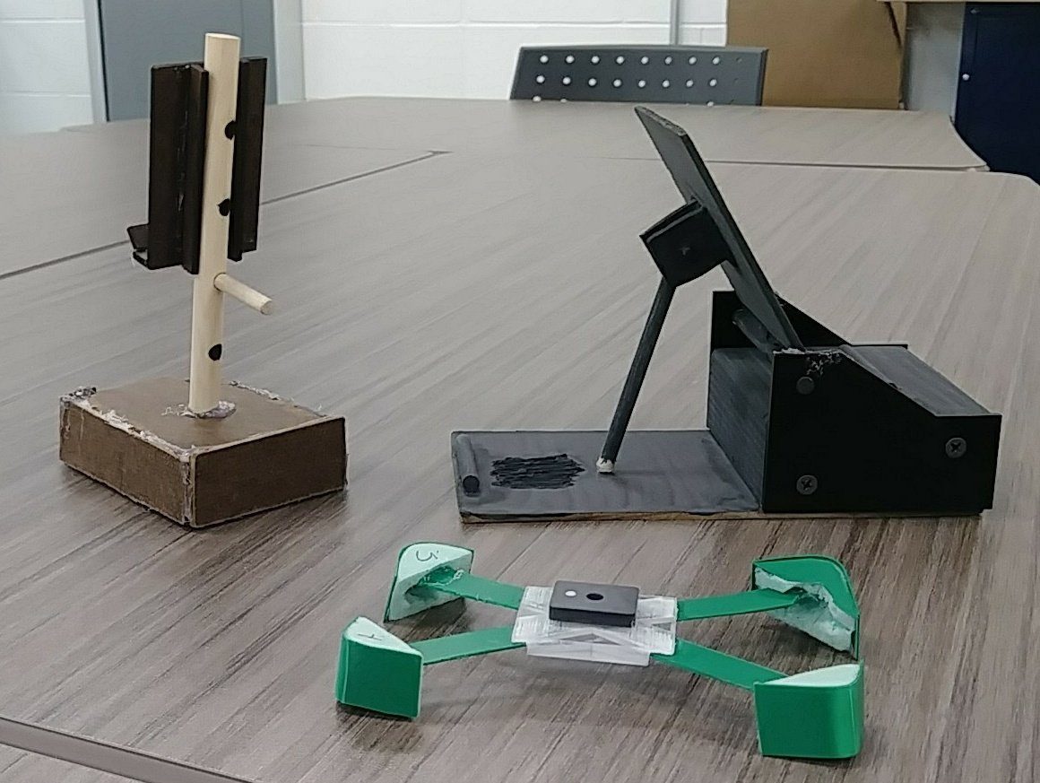 Three different student phone stand designs on a table, made from wood, CNC-cut material, and 3D-printed plastic
