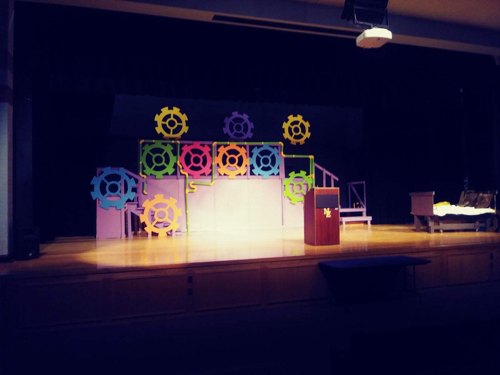 Wide view of the Willy Wonka stage set with the gear wall, lectern, and Grandpa Joe's bed